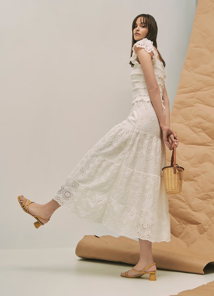 A model wearing a maxi white dress with strappy cylindrical heel sandals and rattan cylindrical crossbody bag.